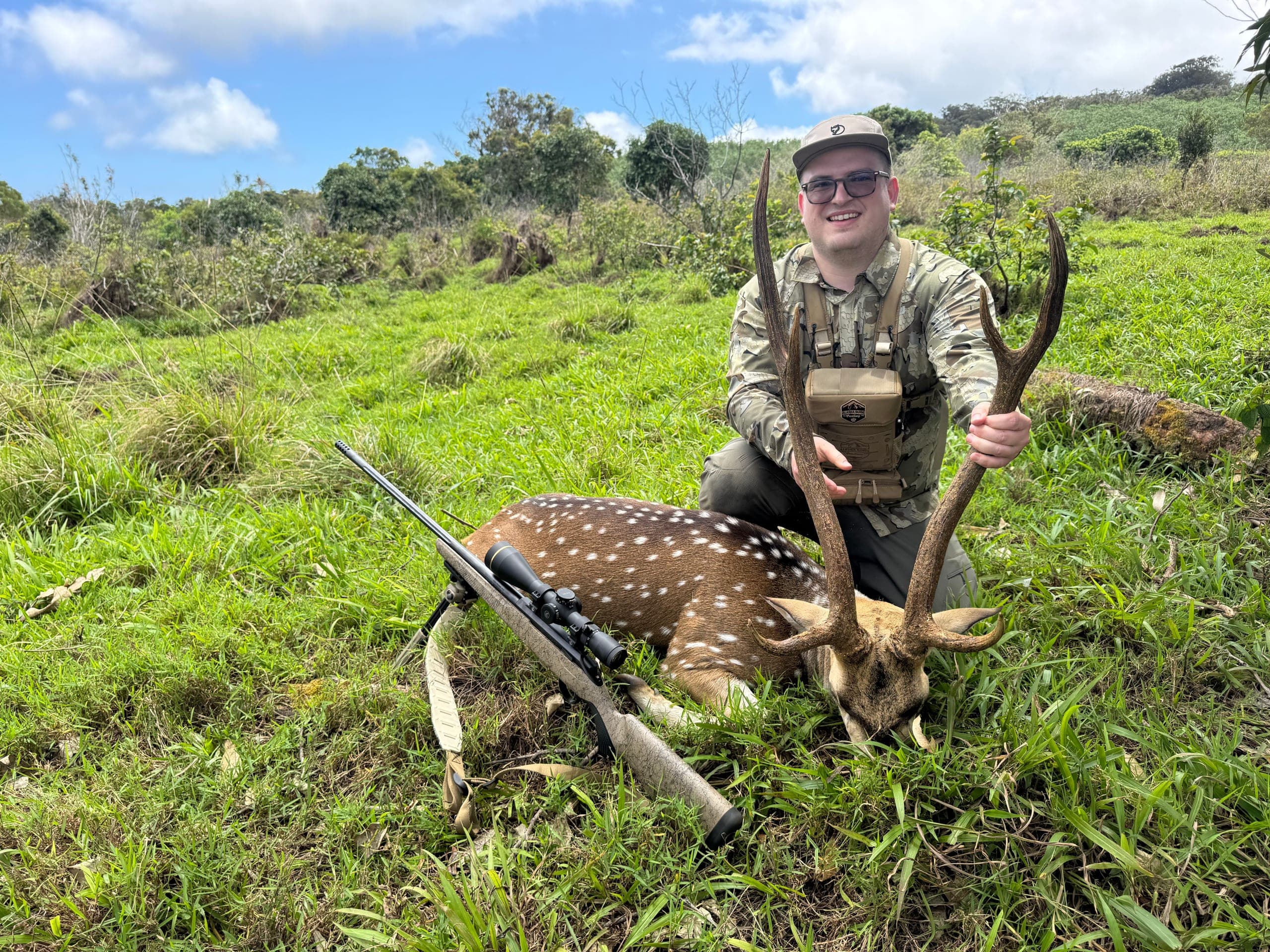 A hunter in camouflage kneels behind a spotted axis buck in bright green pasture under a blue Maui sky